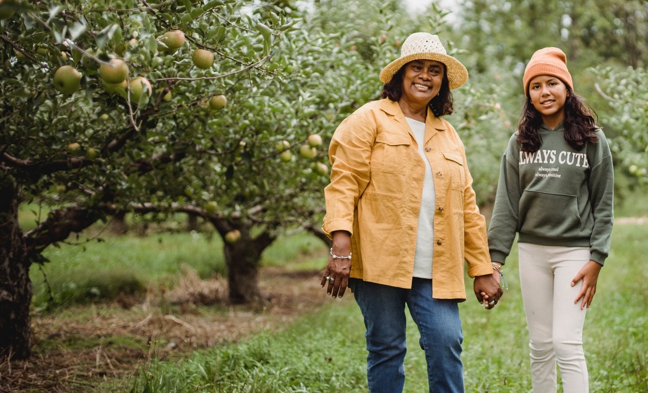 A grandmother and her Autistic grandchild are standing in an apple orchard holding hands.