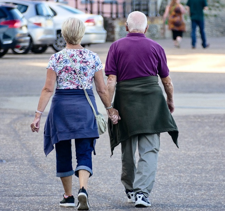 An older neurodiverse couple, an Autistic woman and a typical man, walking and holding hands in a parking lot.